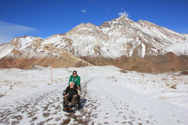 Gids Mariano duwt de rolstoel van Jozef door de sneeuw. Op de achtergrond, besneeuwde bergtoppen.