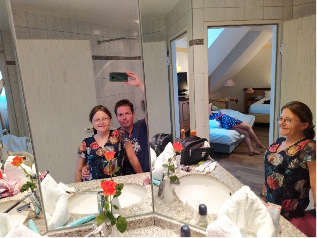 De ouders fotograferen zichzelf in de spiegel van de badkamer. Op de achtergrond ligt de dochter op een bed.