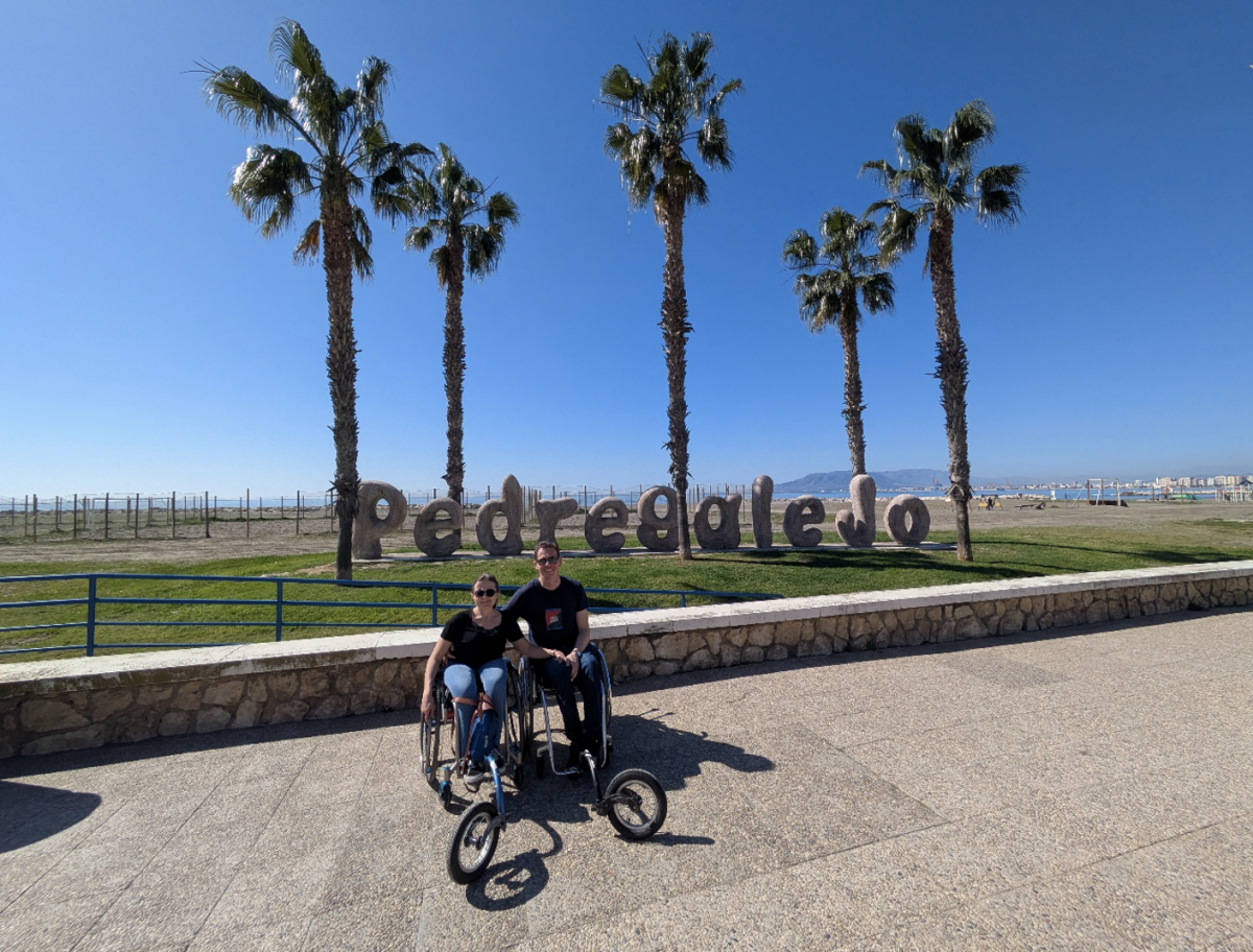 Lien en Thomas, elk in hun rolstoel met "vijfde wiel" poseren bij de letters "Pedregalejo". De hemel is staalblauw.