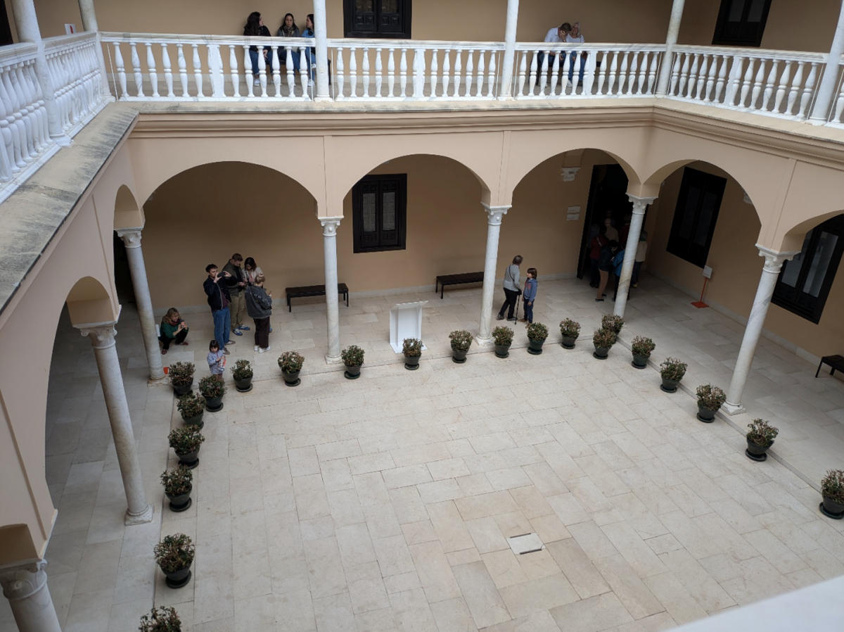 We kijken vanop de eerste verdieping neer over een binnenplaats met typische rondbogen en zuilen. Beneden staan bezoekers in de rij.