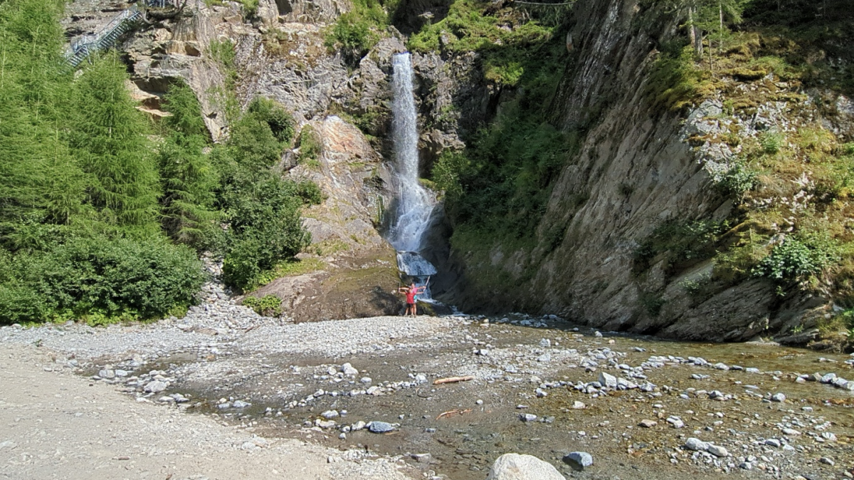 Zoon en dochter poseren met de armen wijd voor de waterval. De weg ernaartoe is bezaaid met stenen en grotere stukken rots. 